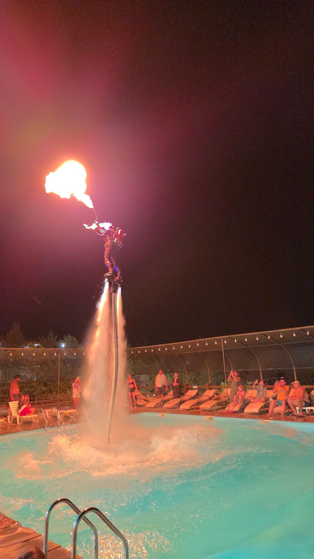 Wide view of a flyboard fire show over a lit hotel pool with guests watching